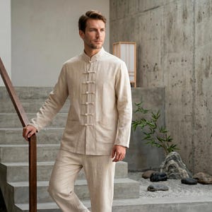 May include: A beige linen two-piece outfit. The long-sleeved shirt has a mandarin collar and frog closures. The matching pants complete the ensemble. The outfit is suitable for casual or formal wear. The model is standing on a set of concrete stairs.