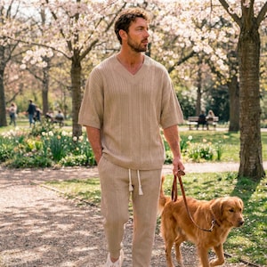 Könnte beinhalten: Beige Strick-Set, bestehend aus einem kurzärmeligen V-Ausschnitt-Pullover und einer Hose mit Kordelzug. Eine Person geht mit einem Golden Retriever-Hund auf einem Weg in einem Park spazieren. Der Hund ist an einer braunen Lederleine.