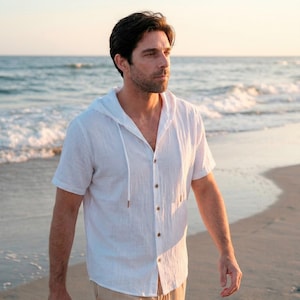 Puede incluir: Camisa blanca con capucha y manga corta, con botones en la parte delantera. La camisa tiene un cordón en la capucha y está hecha de una tela ligera. La persona camina por una playa de arena cerca del mar.