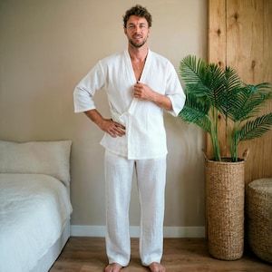 May include: A white linen kimono-style top with matching pants. The top has a wrap-around design with a waist tie and three-quarter sleeves. The pants are loose-fitting. The outfit is worn by a person standing in a room with a bed and a potted plant.