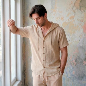 Op de afbeelding: Een beige linnen shirt met capuchon, korte mouwen en een knoopsluiting. Het shirt heeft een trekkoord bij de capuchon en wordt gecombineerd met een bijpassende beige broek. De outfit is casual en comfortabel.