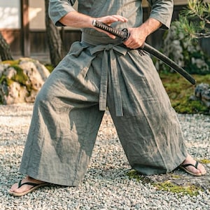 Può includere: Un hakama in lino verde salvia con silhouette a gamba larga e vita annodata, abbinato a un top a maniche lunghe coordinato. L'indumento presenta una spada katana tenuta orizzontalmente all'altezza della vita, con l'impugnatura nera ben visibile.
