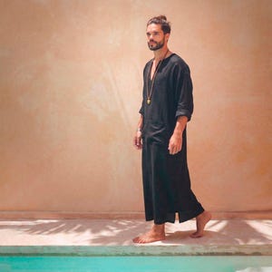 May include: A black linen robe with a V-neck and front pockets. The robe is ankle-length and has three-quarter sleeves. A gold-colored necklace is worn with the robe. The background is a light orange wall and a pool of water.