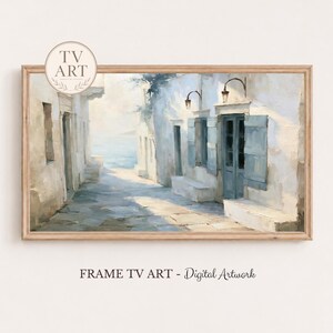 May include: A rectangular digital artwork print in a wooden frame depicting a coastal Mediterranean street scene with white buildings, blue shutters, and two wall-mounted lamps. The text TV ART is visible in a circular badge at the top left.