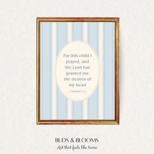 May include: A framed print with a gold-colored frame. The artwork features a light blue and white striped background with an oval-shaped text box. The text reads, "For this child I prayed, and the Lord has granted me the desires of my heart 1 Samuel 1:27." The bottom of the frame reads "BUDS & BLOOMS Art that feels like home."