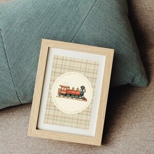 May include: A framed print featuring a vintage train illustration. The train is red and black, set against a cream-colored circle on a plaid background. The frame is made of light-colored wood, and the print is resting against a teal pillow.