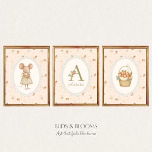 Op de afbeelding: Drie ingelijste kunstprints met een grillig thema. De prints tonen een muis, een bloemrijke initiaal "A" met de naam "Aurora" en een mand met aardbeien. De achtergrond is licht perzikkleurig met aardbeienpatronen. De frames zijn warmbruin.