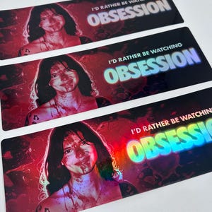 May include: Three rectangular stickers featuring a portrait of a person with dark hair and blood-like splatters on their face and chest. The text I'D RATHER BE WATCHING OBSESSION is printed in white and blue, with a red and black background.