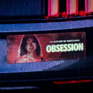 May include: A rectangular bumper sticker featuring a red-toned portrait of a person and the white text I'D RATHER BE WATCHING OBSESSION. The sticker is applied to the dark rear bumper of a vehicle under rainy conditions with red taillights visible.