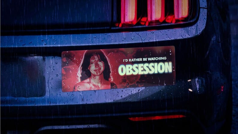 May include: A rectangular bumper sticker featuring a red-toned portrait of a person and the white text I'D RATHER BE WATCHING OBSESSION. The sticker is applied to the dark rear bumper of a vehicle under rainy conditions with red taillights visible.