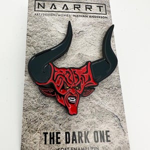 Puede incluir: Un pin de esmalte rojo y negro con un demonio con cuernos. El pin est&aacute; en una tarjeta con el texto "The Dark One" y "Soft Enamel Pin".