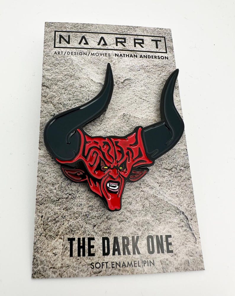 Puede incluir: Un pin de esmalte rojo y negro con un demonio con cuernos. El pin est&aacute; en una tarjeta con el texto "The Dark One" y "Soft Enamel Pin".