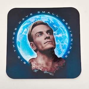 May include: Square coaster featuring a portrait of a man with blonde hair, set against a blue background with a glowing moon. The text "BIG THINGS HAVE SMALL BEGINNINGS" is printed around the edge.