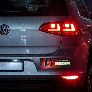 May include: A rectangular bumper sticker applied to the rear of a silver Volkswagen hatchback. The sticker features a graphic of a person with dark hair and the text I'D RATHER BE WATCHING OBSESSION in bold yellow and white lettering.