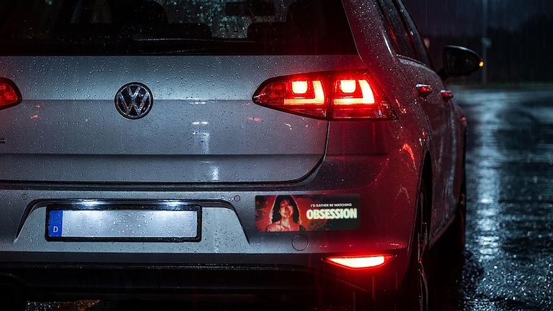 May include: A rectangular bumper sticker applied to the rear of a silver Volkswagen hatchback. The sticker features a graphic of a person with dark hair and the text I'D RATHER BE WATCHING OBSESSION in bold yellow and white lettering.