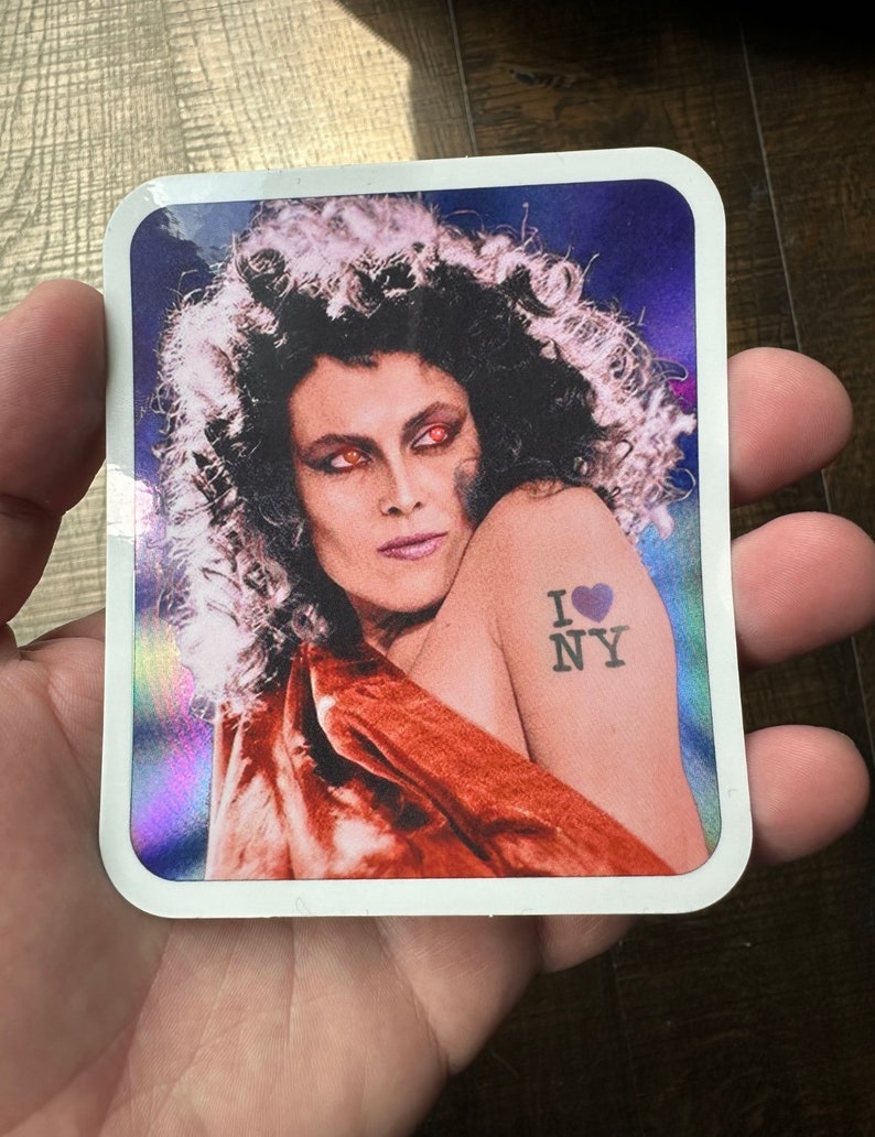 May include: A sticker featuring a woman with dark curly hair and red eyes. She is wearing a red top and has a tattoo on her arm that says "I &hearts; NY".