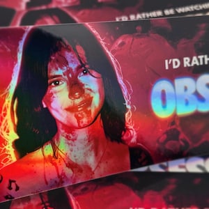 May include: A rectangular sticker featuring a portrait of a person with dark hair, their face covered in red splatters. The text I'D RATHER BE WATCHING OBSESSED is printed in white and holographic blue lettering against a vibrant red and black background.
