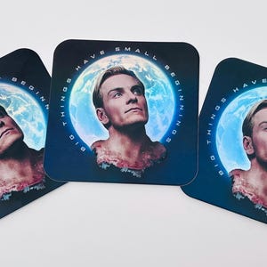 May include: Three square coasters with a portrait of a man against a blue and white planet background. The coasters have the text "BIG THINGS HAVE SMALL BEGINNINGS" around the image. The coasters are black with rounded corners.