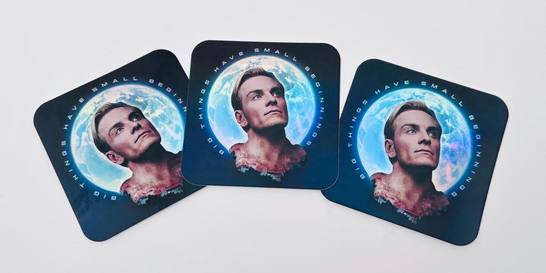 May include: Three square coasters with a portrait of a man against a blue and white planet background. The coasters have the text "BIG THINGS HAVE SMALL BEGINNINGS" around the image. The coasters are black with rounded corners.