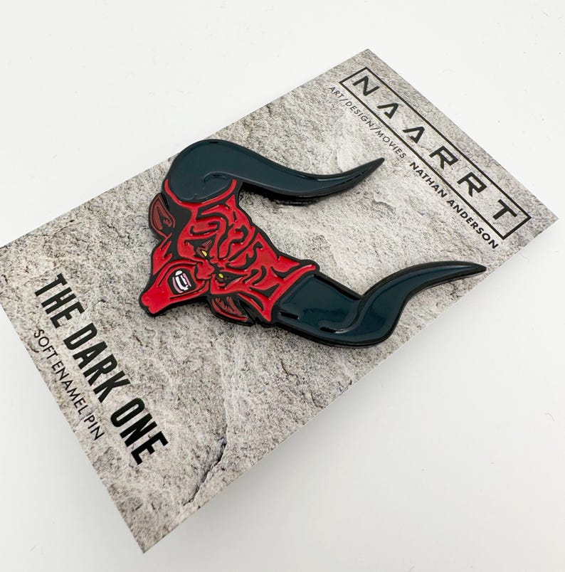 Puede incluir: Un pin de esmalte rojo y negro con una cabeza de demonio con cuernos. El pin dice "The Dark One" y "Soft Enamel Pin". El fondo es una textura de piedra gris. El pin est&aacute; empaquetado en una tarjeta blanca con el texto "NARRT ART/DESIGN/MOVIES NATHAN ANDERSON".