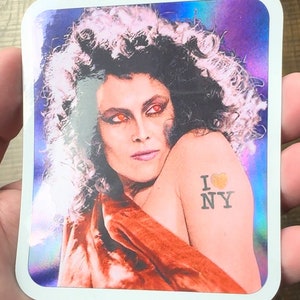 May include: A sticker featuring a woman with long, curly hair and red eyes. She is wearing a red and orange dress and has a tattoo on her arm that says "I love NY" with a heart.