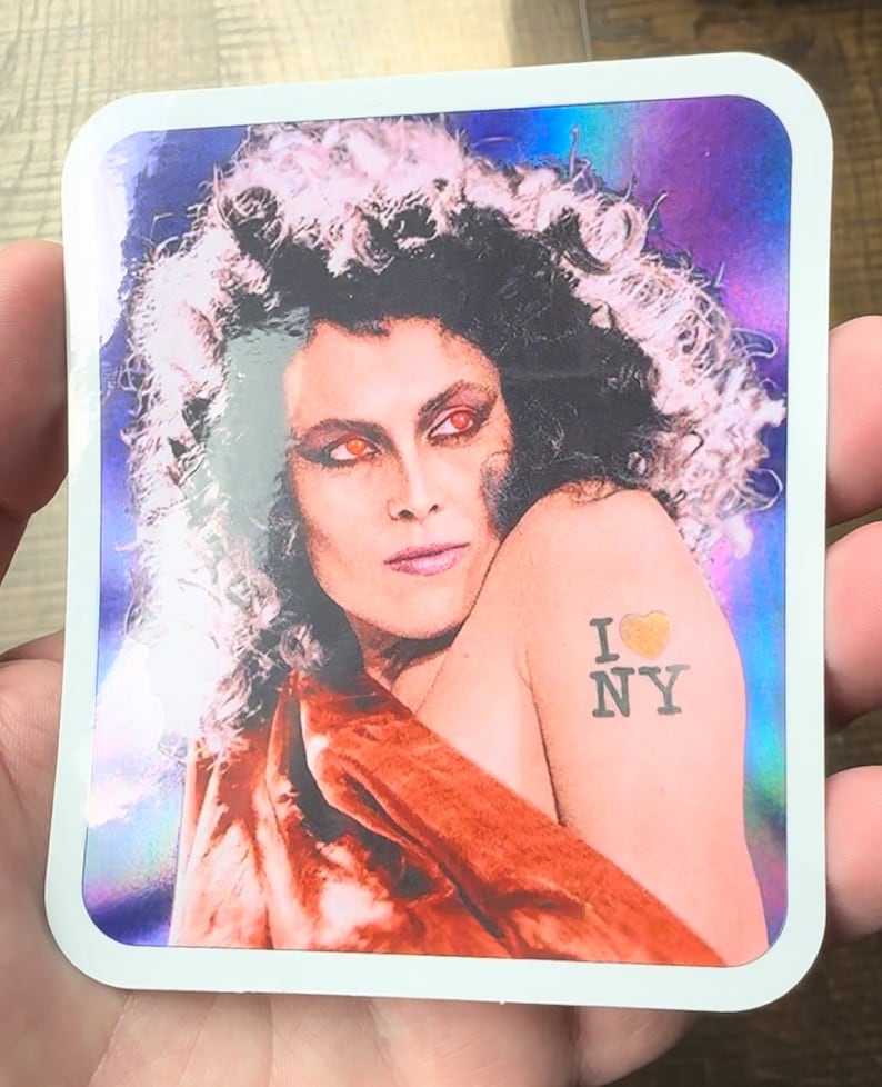 May include: A sticker featuring a woman with long, curly hair and red eyes. She is wearing a red and orange dress and has a tattoo on her arm that says "I love NY" with a heart.