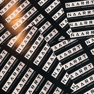 May include: A close-up of a black surface covered with white stickers. The stickers have the text "NAARRT" and "ART/DESIGN/MOVIES NATHAN ANDERSON" repeated multiple times.