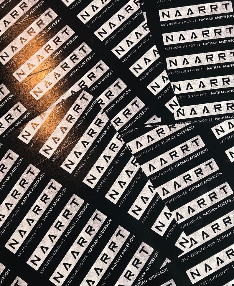May include: A close-up of a black surface covered with white stickers. The stickers have the text "NAARRT" and "ART/DESIGN/MOVIES NATHAN ANDERSON" repeated multiple times.