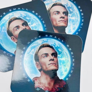 May include: Square coasters with a portrait of a man with blonde hair against a blue background with a planet and the words "HAVE SMALL BEGINNINGS BIG THINGS" in white. The coasters are glossy.