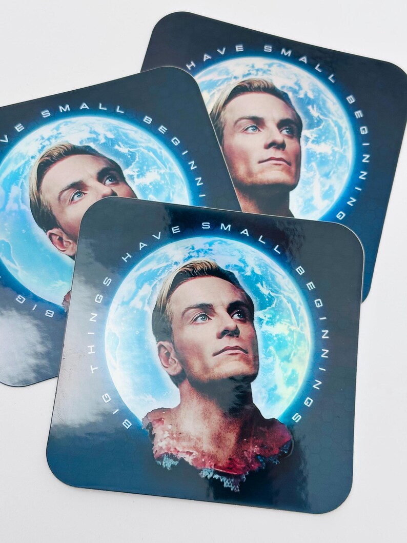 May include: Square coasters with a portrait of a man with blonde hair against a blue background with a planet and the words "HAVE SMALL BEGINNINGS BIG THINGS" in white. The coasters are glossy.