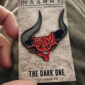 Puede incluir: Un pin de esmalte rojo y negro que representa a un demonio con cuernos. El pin est&aacute; en una tarjeta con el texto "The Dark One" y "Soft Enamel Pin".