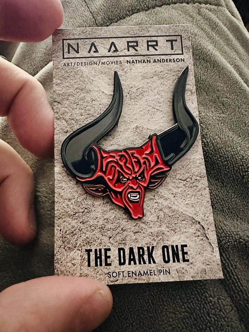 Puede incluir: Un pin de esmalte rojo y negro que representa a un demonio con cuernos. El pin est&aacute; en una tarjeta con el texto "The Dark One" y "Soft Enamel Pin".