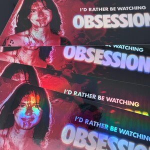 May include: A rectangular sticker featuring a holographic finish with a portrait of a person with dark hair and blood-like splatters on their face. The text I'D RATHER BE WATCHING OBSESSION is printed in white and blue block letters.