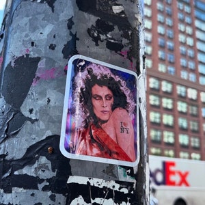 May include: A sticker on a weathered pole with a woman with long curly hair wearing a red shirt and a tattoo that says "I love NY" with a red heart.