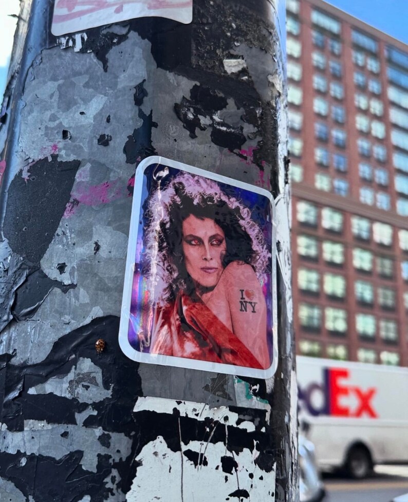 May include: A sticker on a weathered pole with a woman with long curly hair wearing a red shirt and a tattoo that says "I love NY" with a red heart.