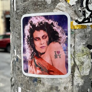 May include: A sticker with a woman with long dark hair and red eyes wearing a red and orange dress. The woman has a tattoo on her shoulder that says "I love NY" with a red heart.