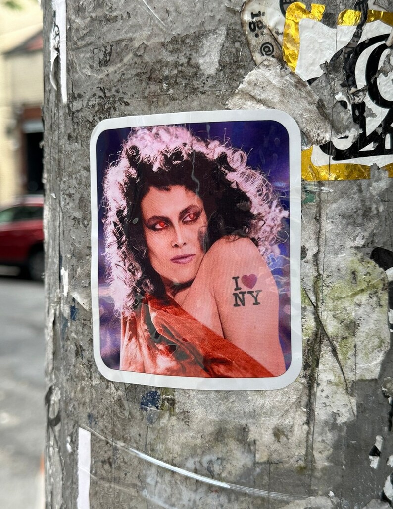 May include: A sticker with a woman with long dark hair and red eyes wearing a red and orange dress. The woman has a tattoo on her shoulder that says "I love NY" with a red heart.