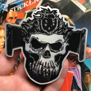 May include: A metallic sticker featuring a skull design with a black and silver color scheme. The skull has detailed features and is set against a brushed metal background. The sticker has a unique, edgy aesthetic.
