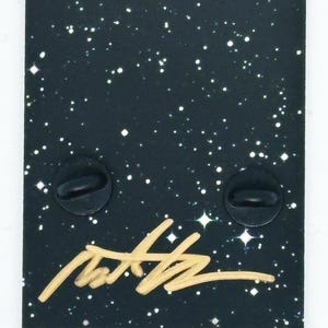 Puede incluir: Una tarjeta negra con un fondo de purpurina dorada y texto blanco que dice "www.naarrt.com". La tarjeta tambi&eacute;n presenta una firma dorada, el texto "Soft Enamel Pin" y "Designed by Naarrt 20/125".