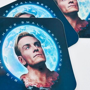 May include: Square coasters featuring a man with blonde hair and blue eyes, set against a blue planet backdrop. The coasters have the text "HAVE SMALL BEGINNINGS" around the planet image. The coasters are dark blue.