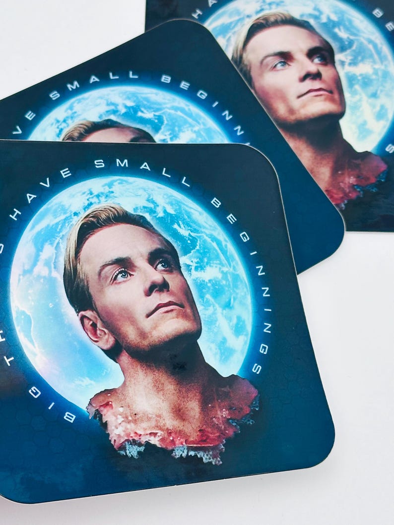 May include: Square coasters featuring a man with blonde hair and blue eyes, set against a blue planet backdrop. The coasters have the text "HAVE SMALL BEGINNINGS" around the planet image. The coasters are dark blue.