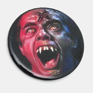 May include: Round button with a horror-themed illustration. The image features a screaming face, half red and half blue, with sharp fangs and glowing yellow eyes. The button has a glossy finish and a black border.