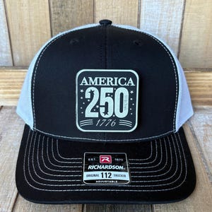 May include: A black and white trucker hat featuring a square patch on the front with the text AMERICA 250 and 1776. The hat has a black front panel, white mesh back panels, and a black brim with white stitching. A Richardson 112 label is on the brim.