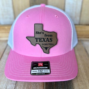 May include: A pink and white trucker hat featuring a brown leather-like patch shaped like the state of Texas. The patch is engraved with the text She's from TEXAS I CAN TELL and a longhorn skull icon. A black Richardson 112 sticker is on the brim.