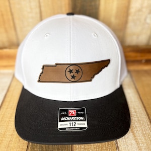 May include: White and black trucker hat with a brown leather patch featuring the outline of Tennessee and three stars. The hat has a curved brim and an adjustable strap. The patch reads "Tennessee".