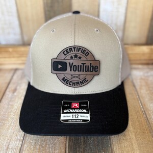 May include: A tan and black Richardson 112 trucker hat with a white mesh back. The hat features a circular patch with the words "CERTIFIED YOUTUBE MECHANIC" and the YouTube logo.