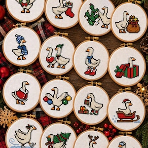 May include: A collection of fifteen circular wooden embroidery hoops, each containing a white fabric panel with a unique cross-stitch design of a white goose engaged in various holiday and winter activities, including wearing scarves, hats, and interacting with gifts.