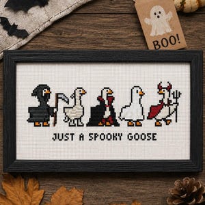May include: A framed cross-stitch embroidery featuring five pixel-art geese dressed in Halloween costumes, including a grim reaper, a vampire, and a devil, with the text "JUST A SPOOKY GOOSE" stitched in black thread on a white fabric background.