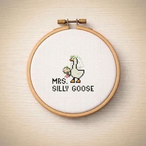 Pode incluir: Bordado em ponto cruz num bastidor de madeira com um ganso pixelizado a usar véu e a segurar flores. O texto "MRS. SILLY GOOSE" está bordado por baixo. O bordado está sobre um fundo claro com grão de madeira.