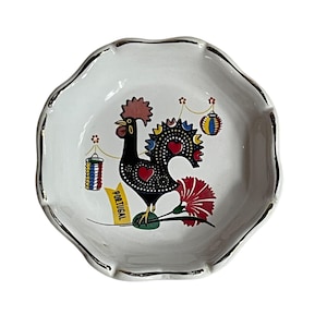 May include: White ceramic decorative plate with a scalloped edge and gold trim. The plate features a black rooster design with red, yellow, and blue accents, and the word "PORTUGAL" in yellow. The rooster is holding a red flower.